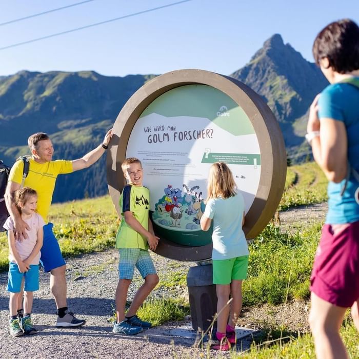 Group of people standing by a large round informational sign with mountains in the background.