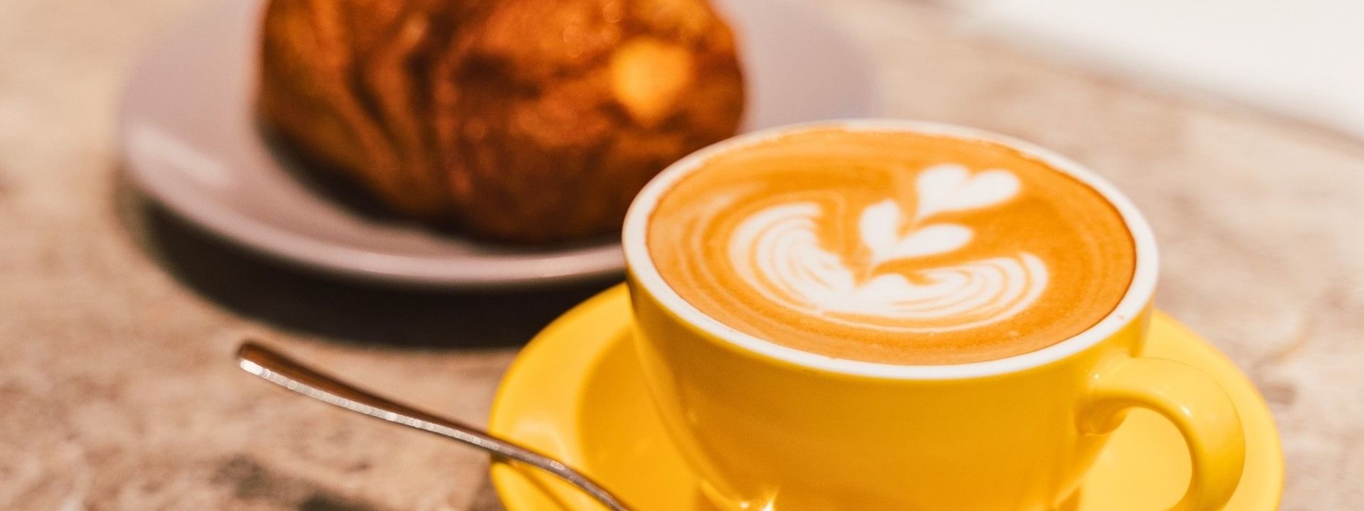 Latte with heart design on yellow cup and spoon, with a croissant on a plate behind.
