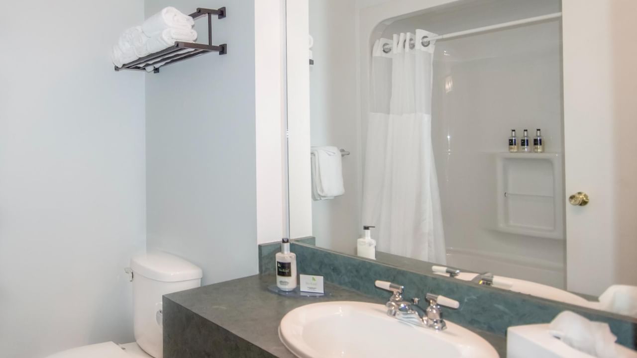 Bathroom with sink, mirror, toilet, and shower stall with folded towels and bottles on shelf.