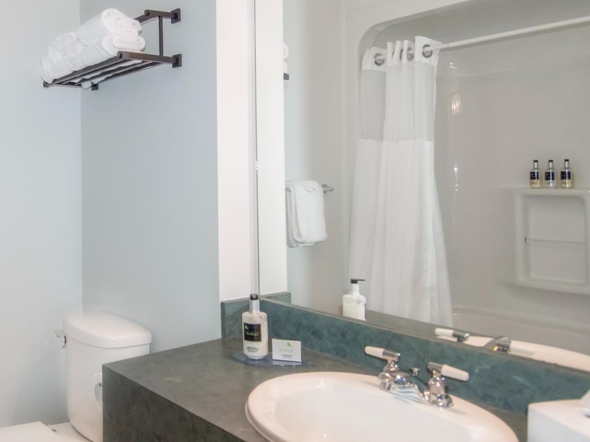 Clean bathroom with white toilet, sink, and mirror reflecting shower with towels and toiletries.