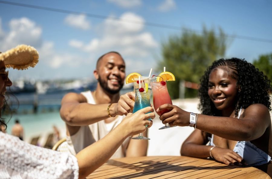 Hog Bar with Friends toasting colorful cocktails by a wooden table at Warwick Paradise Island Bahamas
