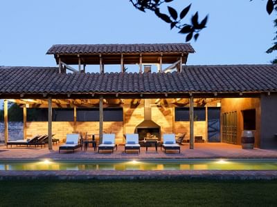 Lounge chairs by a lit pool under a tiled roof pavilion by a fireplace at NOI Blend Colchagua