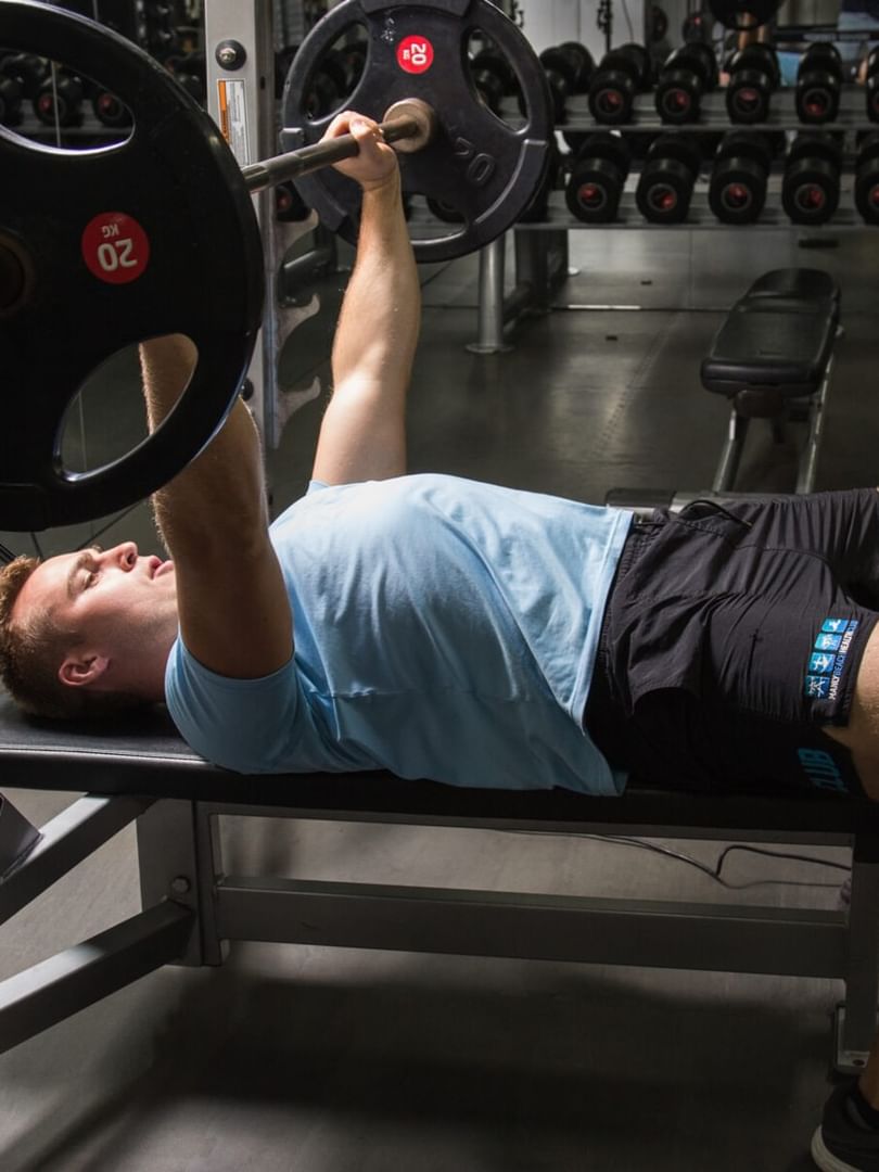Elysium Manly gym features a man pressing a heavy barbell on a workout bench by a dumbbell rack, a featured hotel in Manly