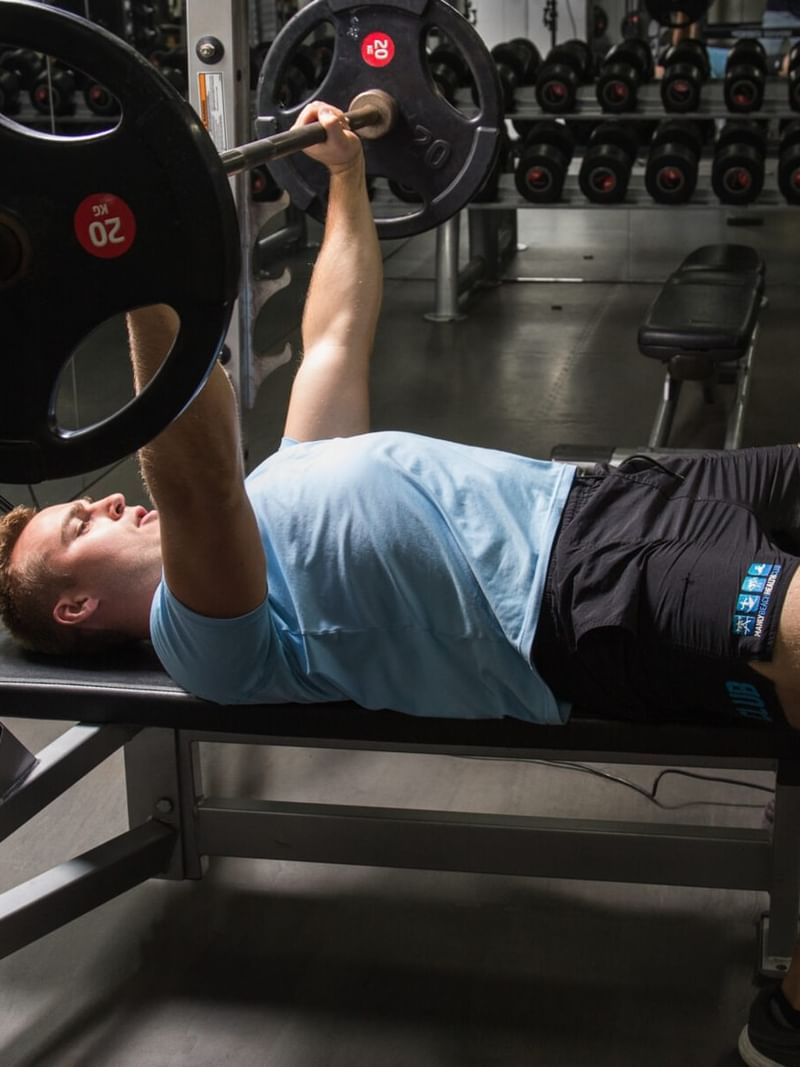 Elysium Manly gym features a man pressing a heavy barbell on a workout bench by a dumbbell rack, a featured hotel in Manly