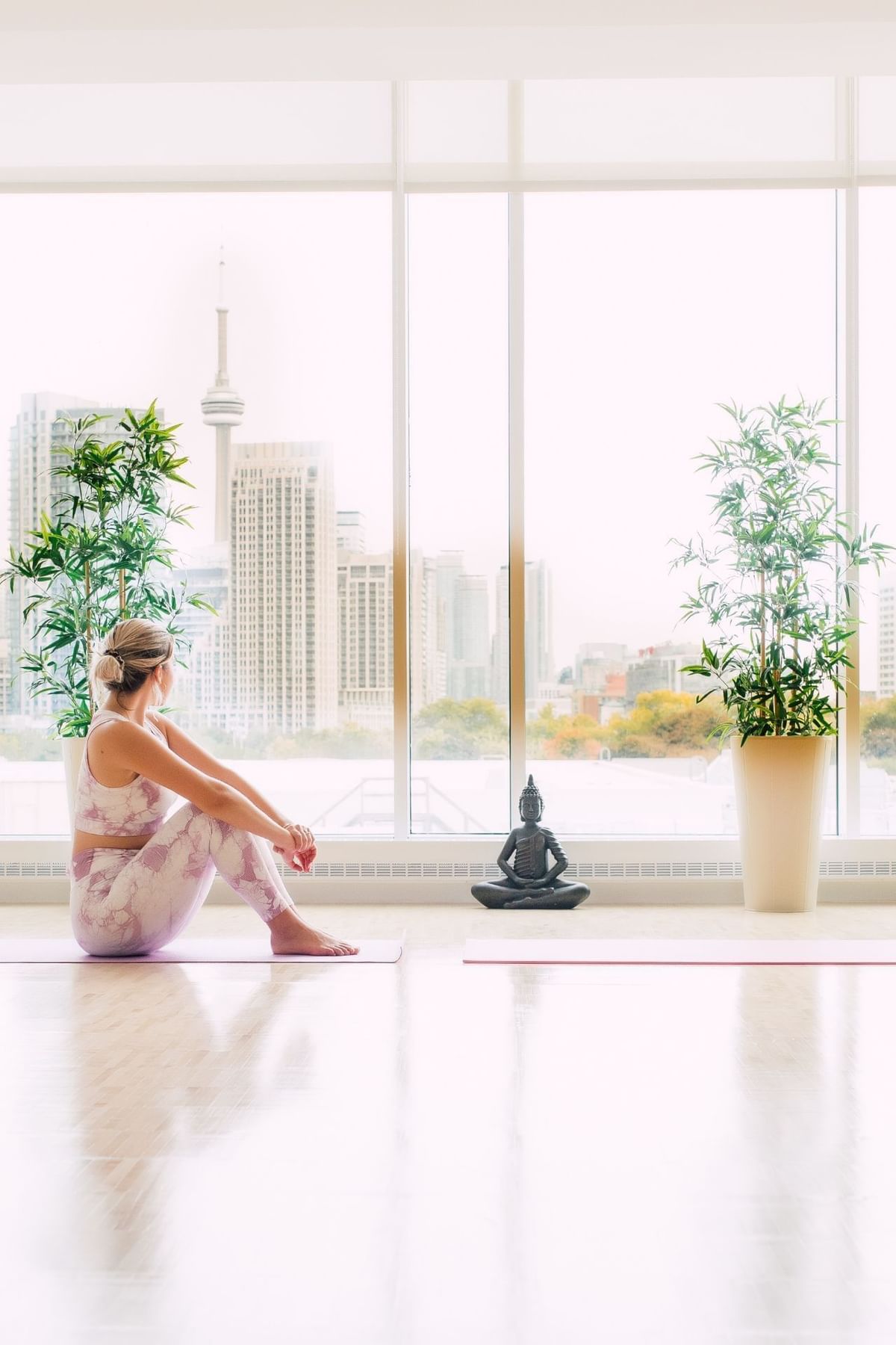 Woman sitting on yoga mat facing floor-to-ceiling windows with potted bamboo plants and city view at Hotel X Toronto
