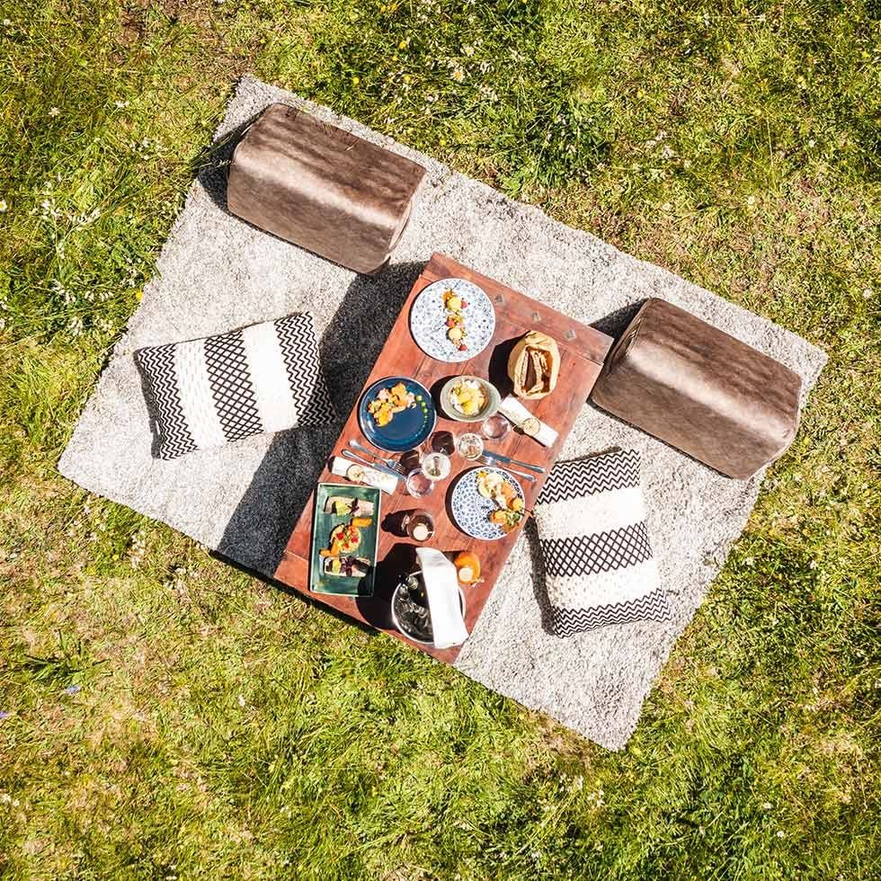 Picnic table with food, plates, and cushions on a gray mat atop grass.