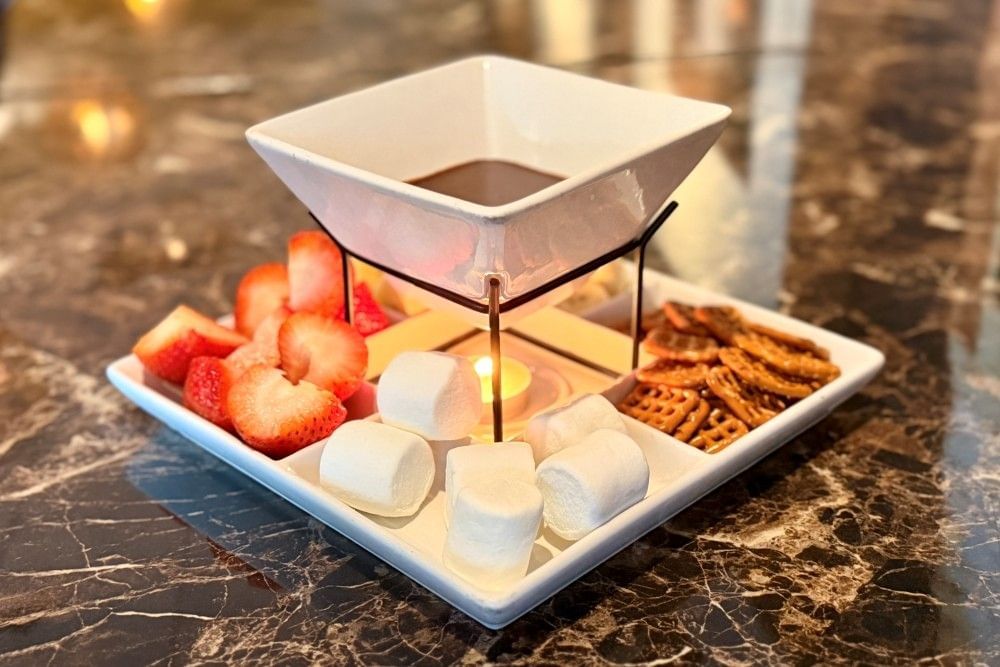 A close-up of a square bowl with chocolate fondue surrounded by marshmallows, pretzels, and strawberries.