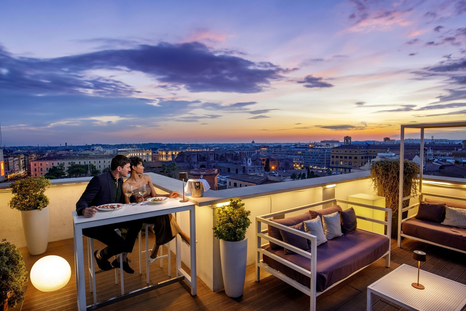 A couple sharing a romantic dinner on a rooftop terrace, overlooking a vibrant city skyline at The Tribune Hotel
