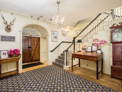 Wooden desk with orchids by a curved stair, under a chandelier near a deer head and oak door at Simsbury 1820 House