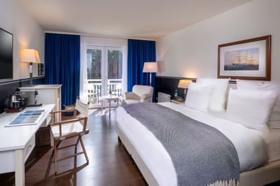 A hotel room with a bed, desk, chair, lamp, and balcony overlooking trees.