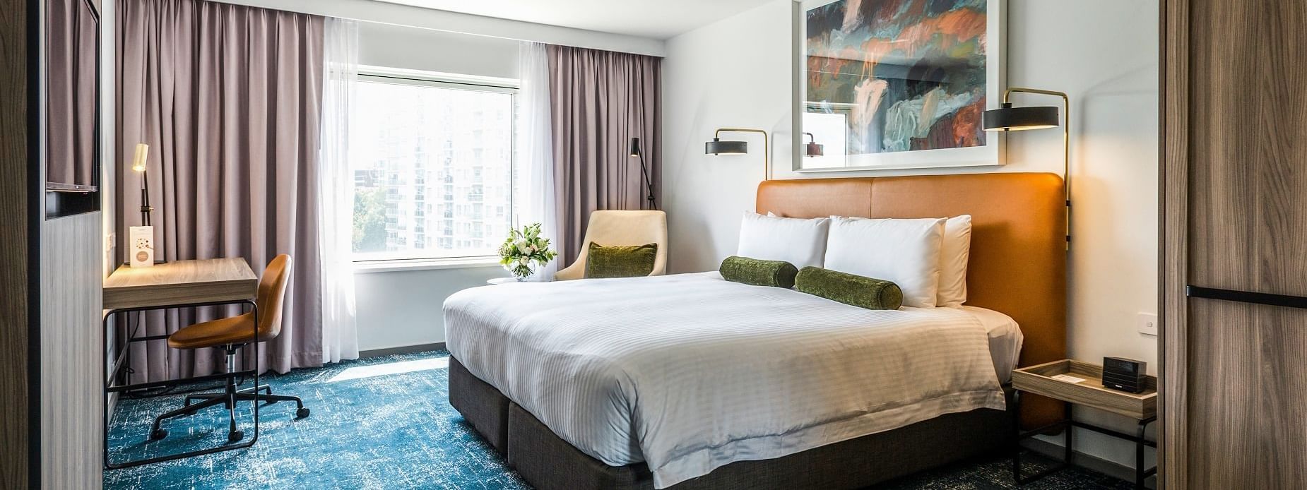 Bed on blue textured carpet near desk with lamp in spacious layout of Deluxe King Room at Novotel Sydney Darling Square