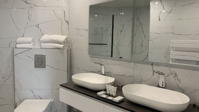 Modern bathroom with two white sinks, marble walls, a toilet, and a large mirror above the vanity.