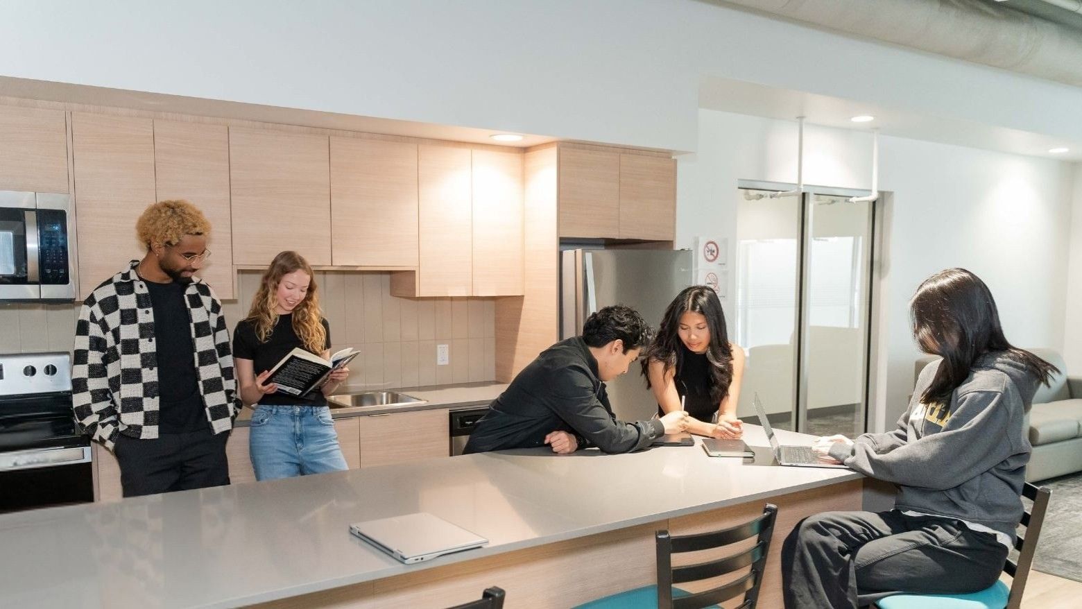 Students studying and socializing in a bright communal kitchen with large island and modern stainless steel appliances.