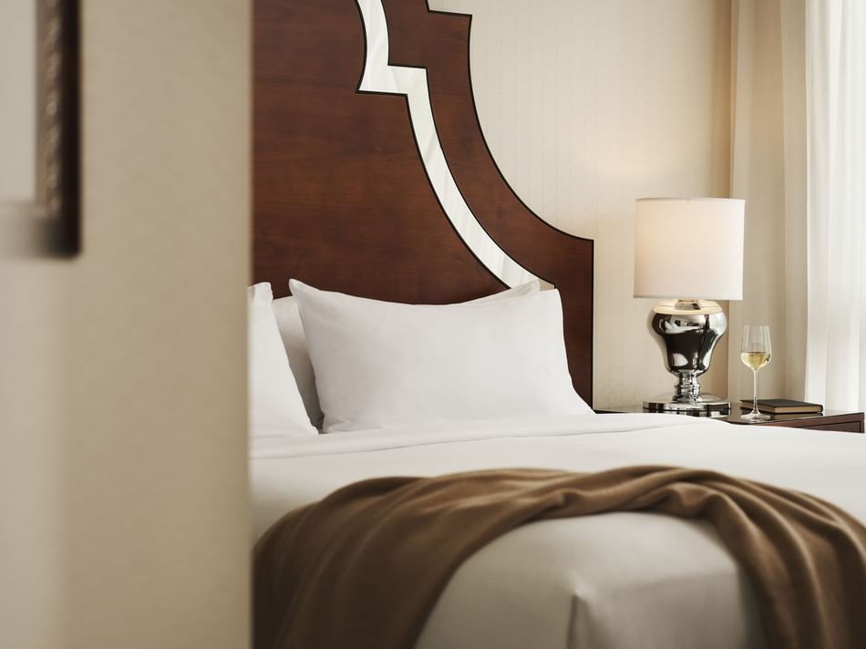 A bedroom with a white bed, brown headboard, white pillows, a lamp, and a wine glass on a table.