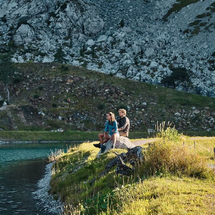 A couple sits on a rock by a lake with rocky mountains in the background.