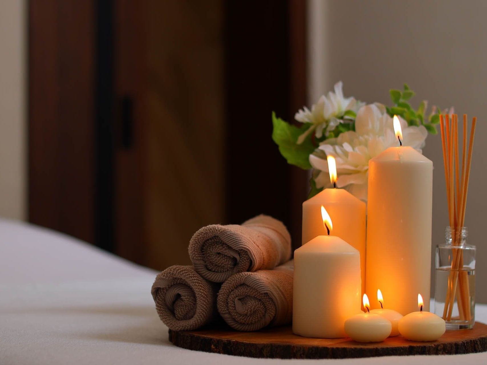 Candles, towels, and floral arrangement on a wooden tray atop a bed in a spa setting.