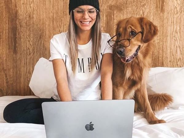 Woman on laptop in bed next to golden retriever with glasses