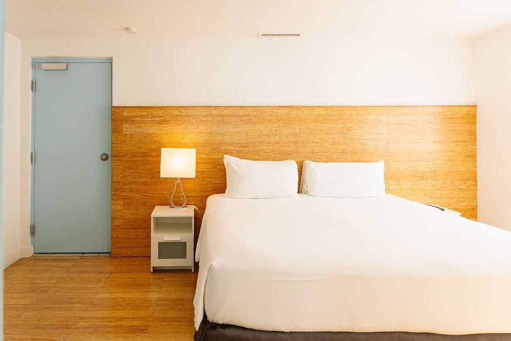 A hotel room with a king-sized bed, wooden floor, wooden headboard, white bedding, blue door, and a nightstand with a lamp.