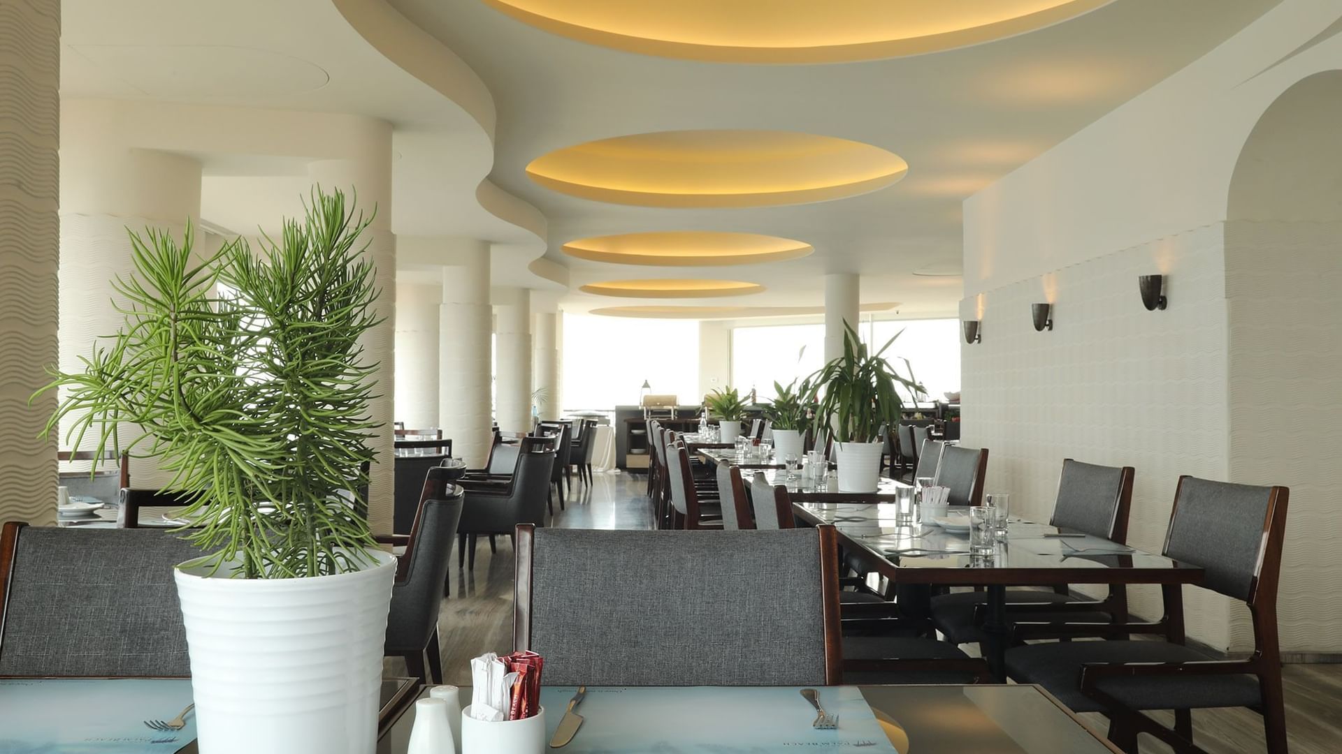 Dining area with plants by tables under round lights, surrounding grey chairs in Lobby Area at Warwick Palm Beach Hotel