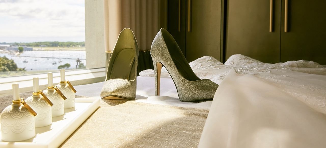Silver high heels on bed with a wedding dress and perfume bottles by window overlooking a lake at Hotel X Toronto