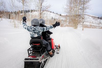 Two excited riders with arms raised gliding through snowy terrain on a snowmobile near Hotel Park City Autograph Collection 