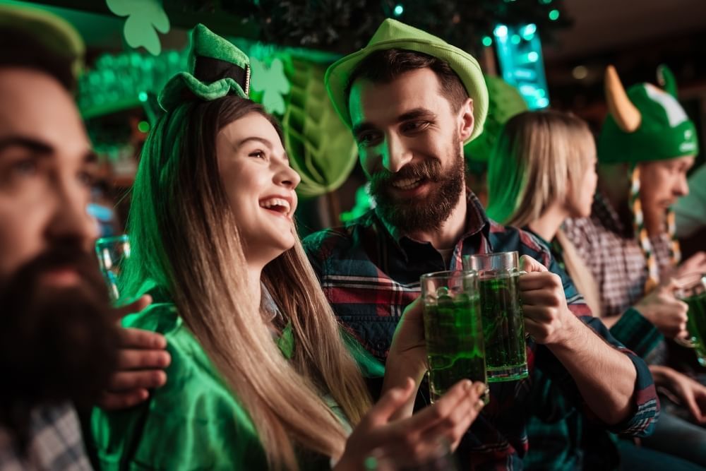 A man and a woman in green hats toast glasses of green beer while smiling in a small crowd on St. Patrick's Day.