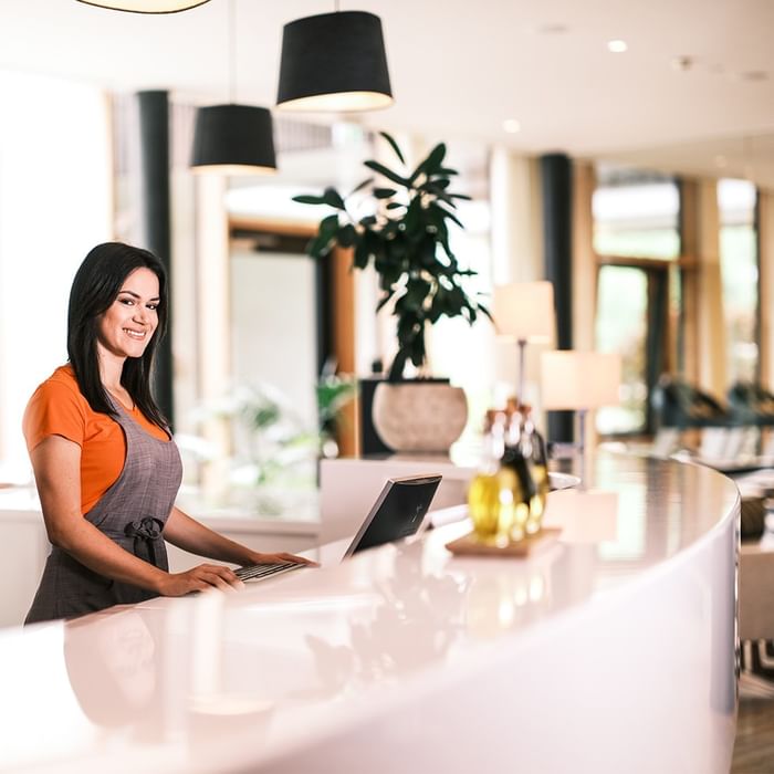 Woman in apron at a reception desk with laptop, smiling, in a modern hotel lobby.