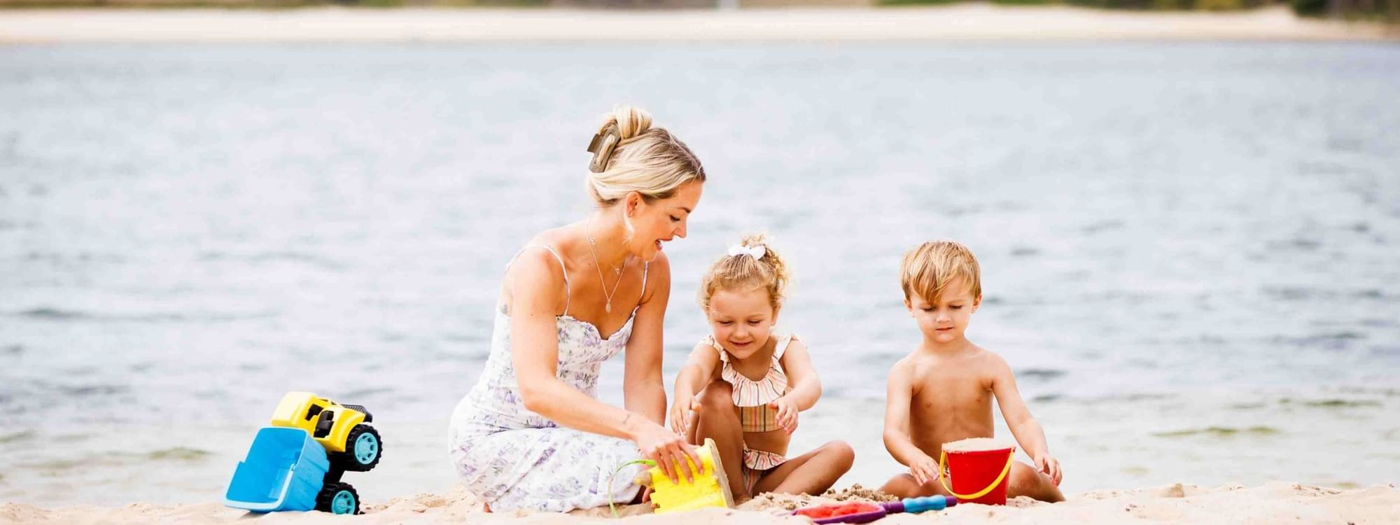 Woman and two children building sandcastles on a sandy beach with toys near Novotel Sunshine Coast Resort