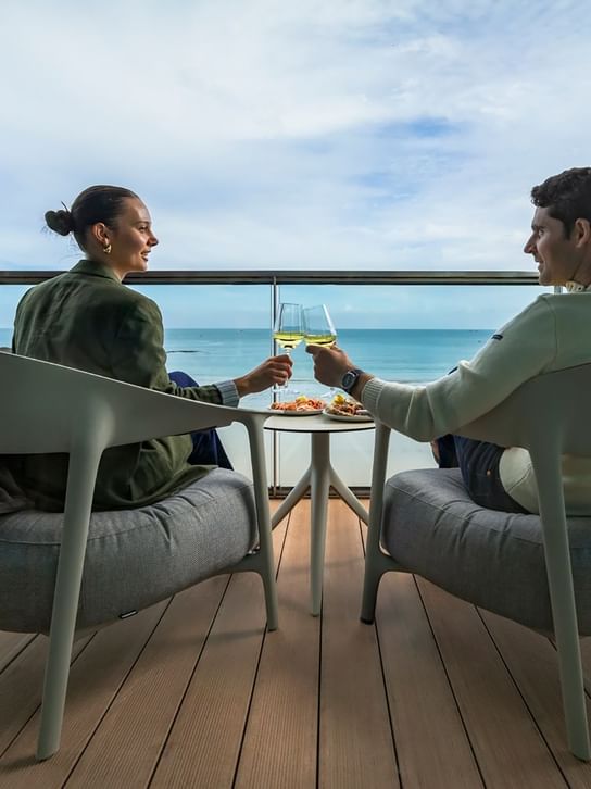 A couple toasts with sparkling wine on a balcony, enjoying ocean views and a shared plate at Oceania Hotels