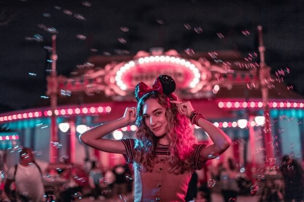 A young woman wearing Minnie ears stands in front of a brightly lit attraction.