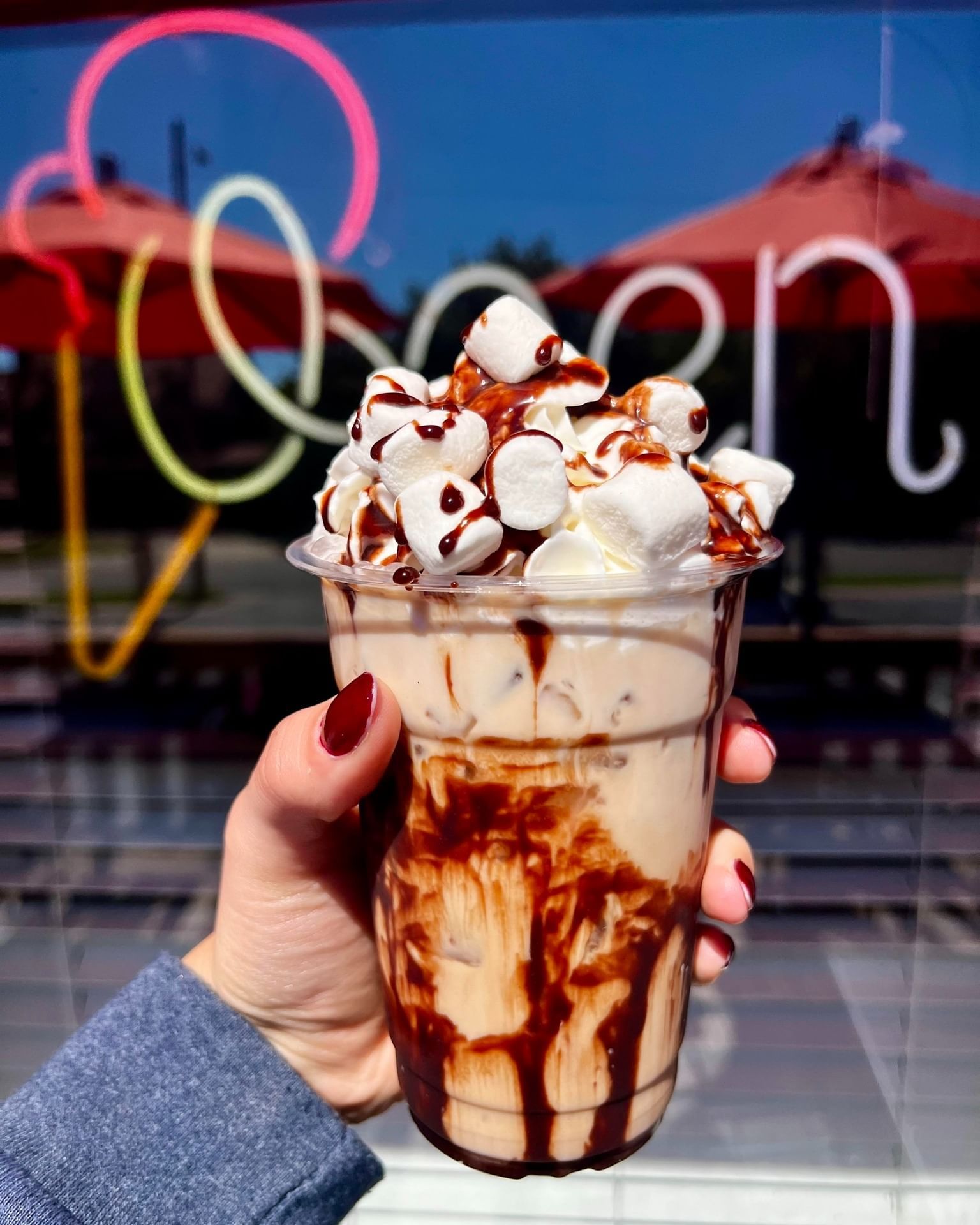 A hand holding a cup of iced coffee topped with marshmallows and chocolate sauce