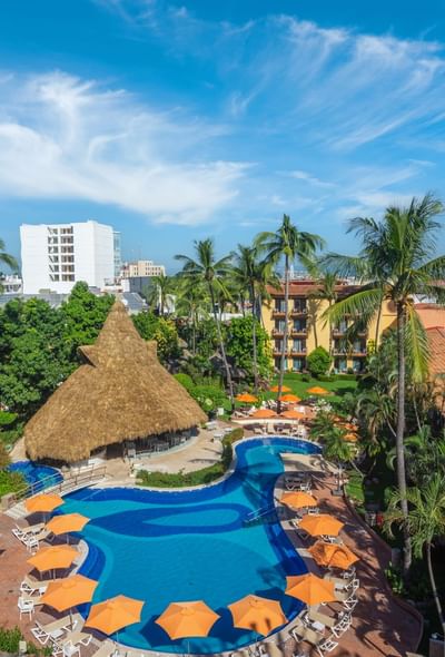 Aerial view of resort with pool, lounge chairs, umbrellas, hut, and yellow building among palm trees.