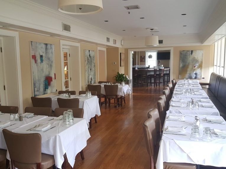 Long dining tables with linens by leather chairs, under abstract art near a bar and windows at Simsbury 1820 House