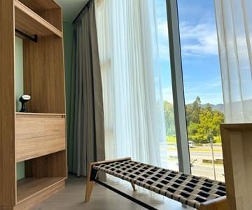 Wooden wardrobe next to woven luggage bench by floor-to-ceiling window overlooking hills at Unika Hotels