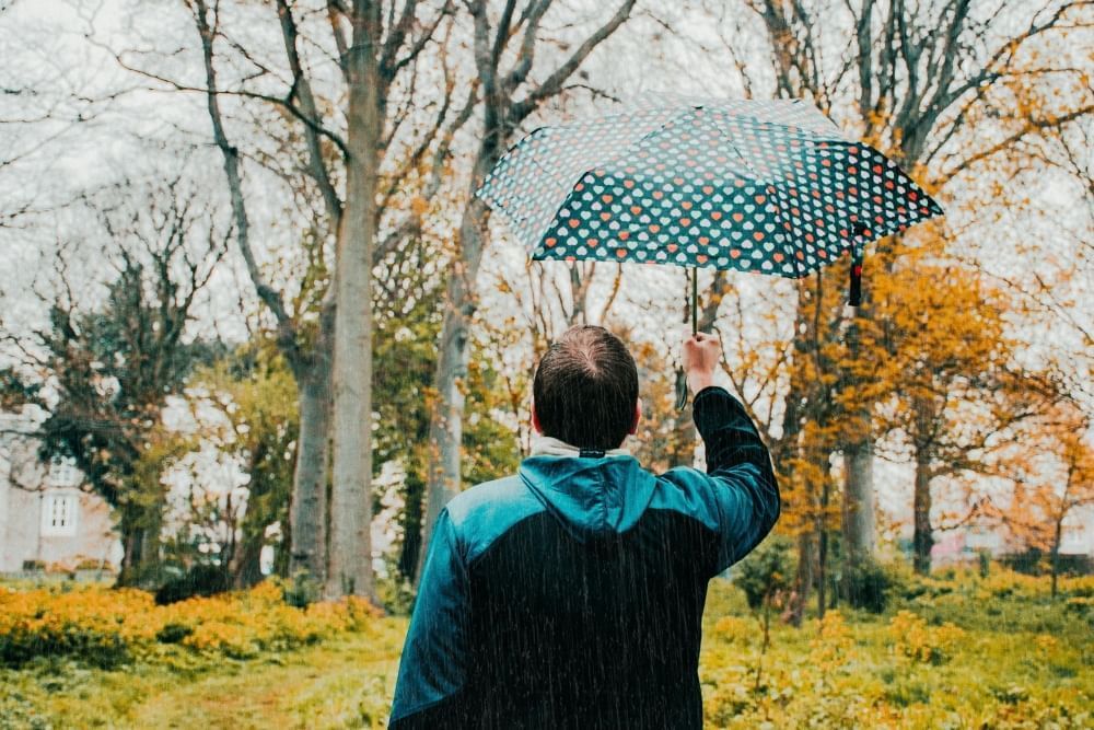 A man wearing a raincoat holding an umbrella in the rain with fall foliage in the background.
