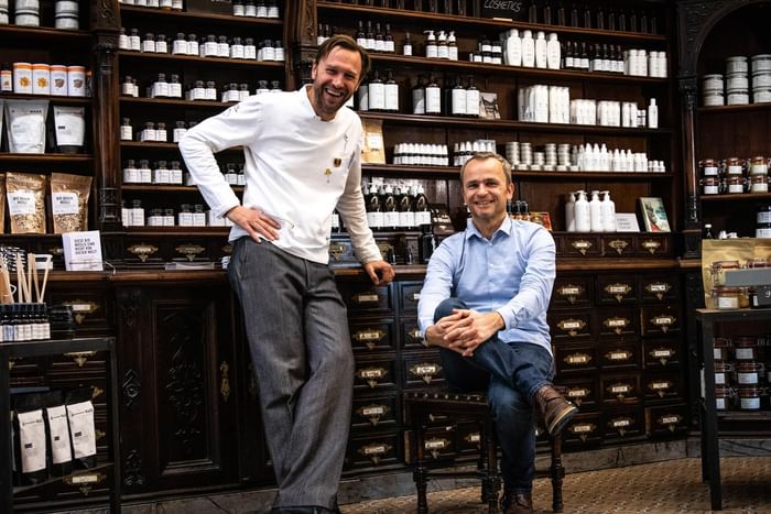 Two men stand and sit in Saint Charles Apothecary, filled with bottles and jars of various products near Hotel Motto Vienna