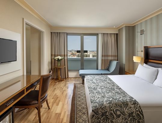 Deluxe Room at Titanic City Taksim with a bed, desk, chair, TV overlooking a cityscape.