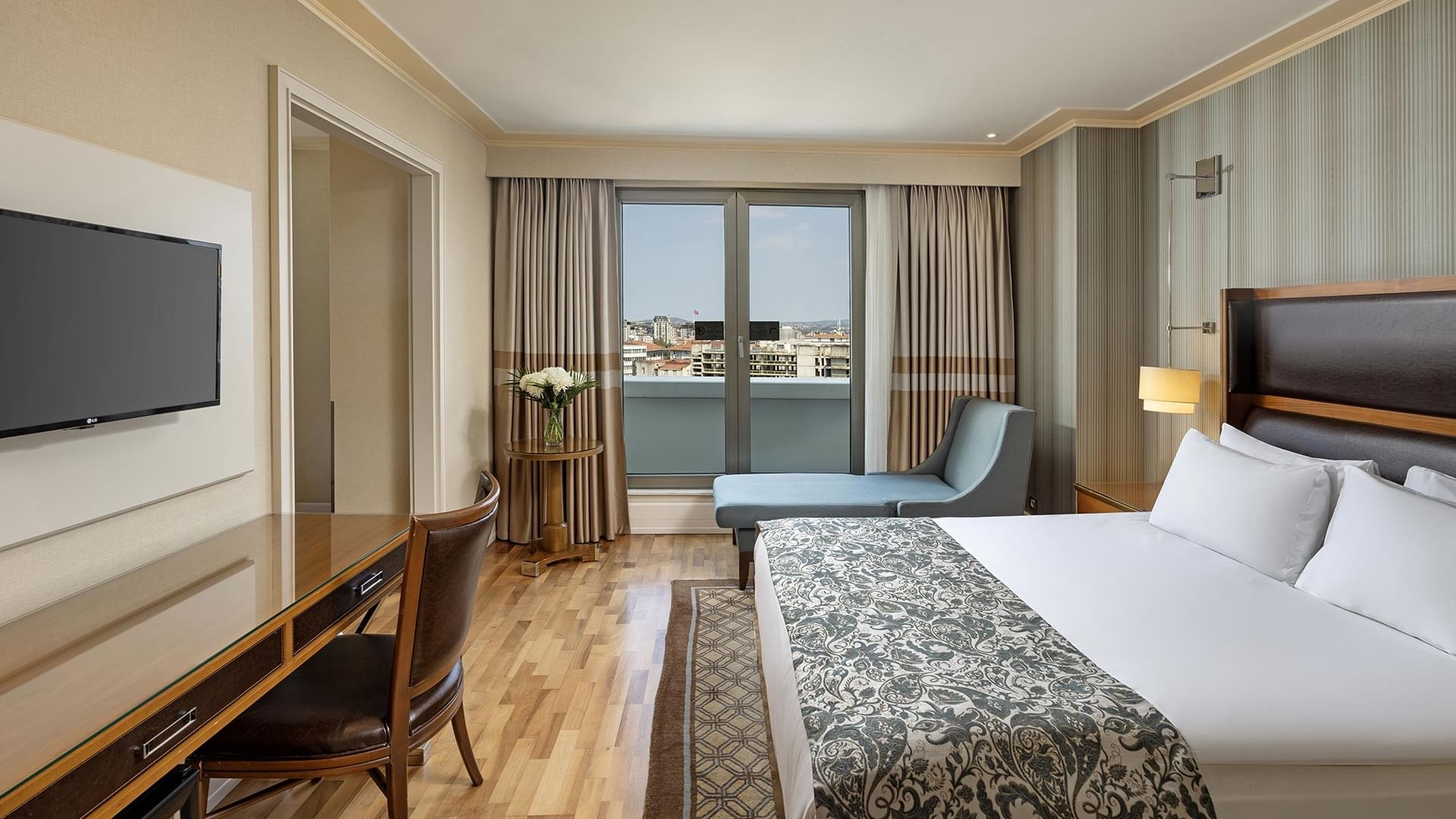 Titanic City Taksim'deki deluxe odanın iç görünümü, büyük yatak, masa, sandalye ve geniş pencere.