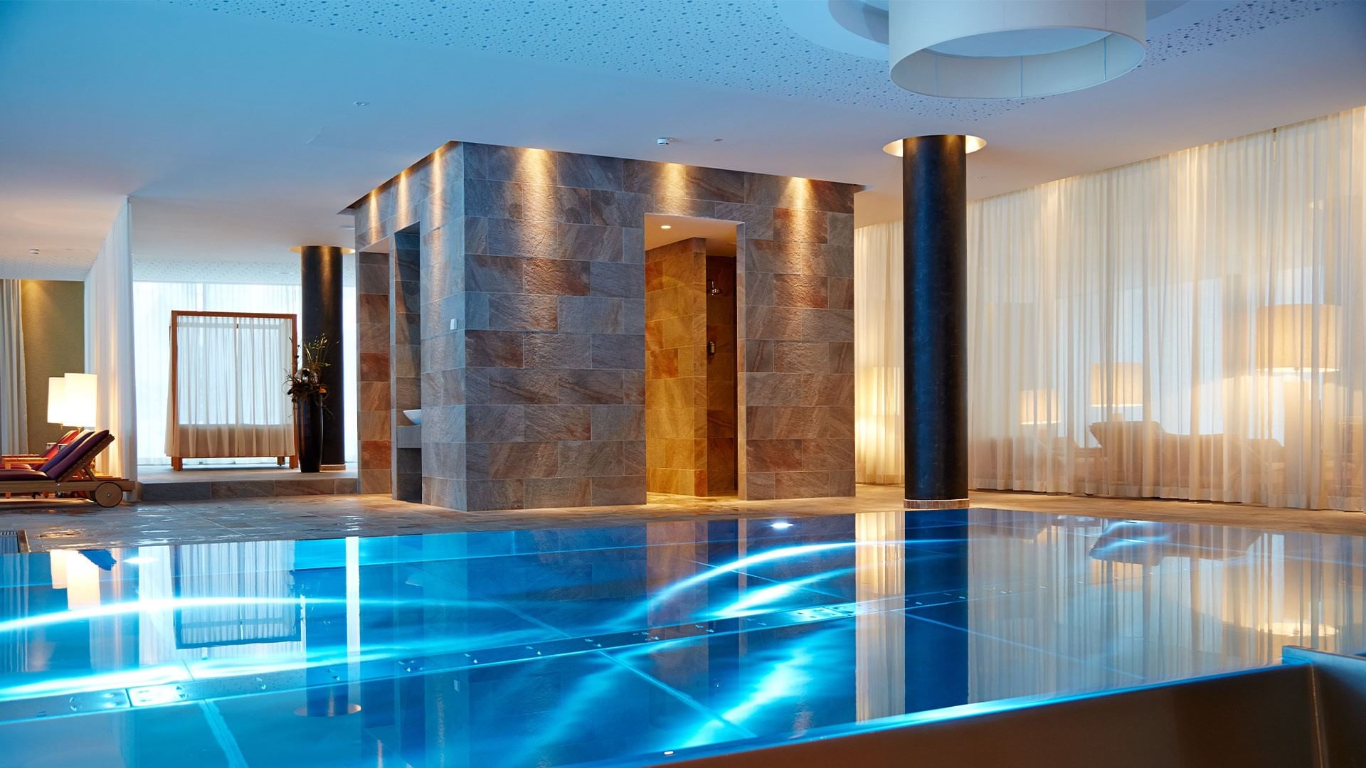 Luxuriöses Spa mit blau beleuchtetem Pool, Steinwänden und eleganten Vorhängen.