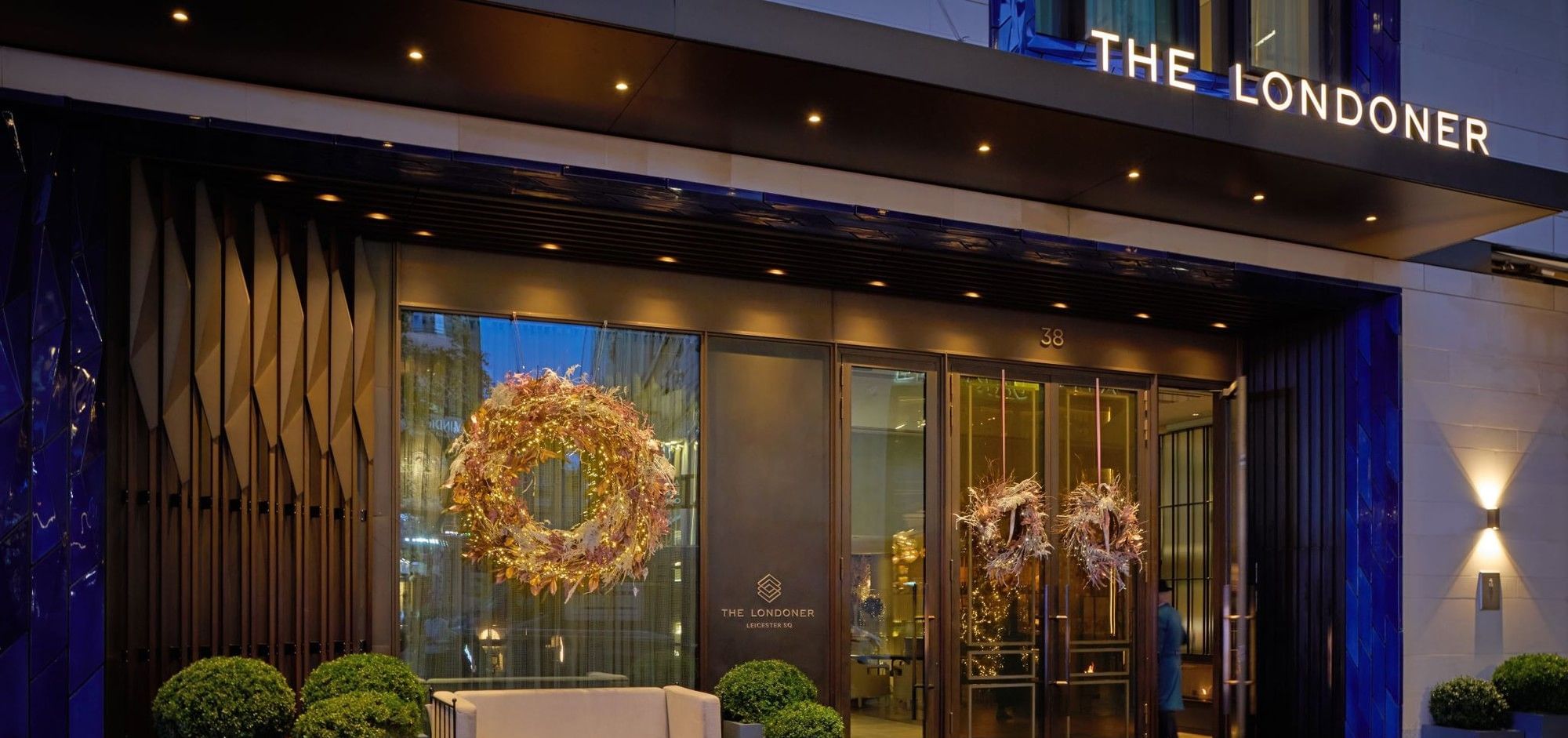 Hotel entrance featuring bright Christmas decorations by windows under the signage at The Londoner Hotel