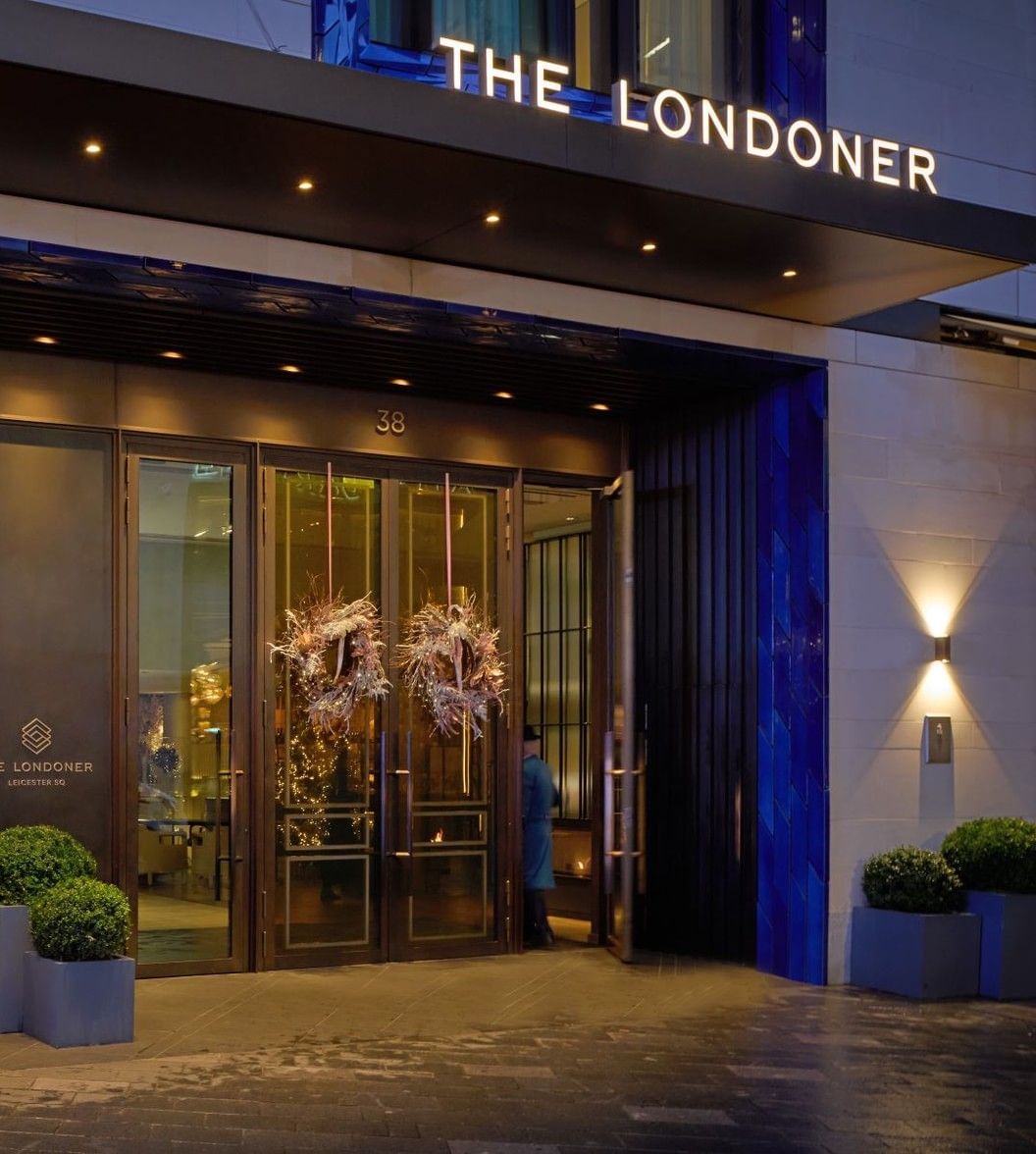 Hotel entrance featuring bright Christmas decorations by windows under the signage at The Londoner Hotel