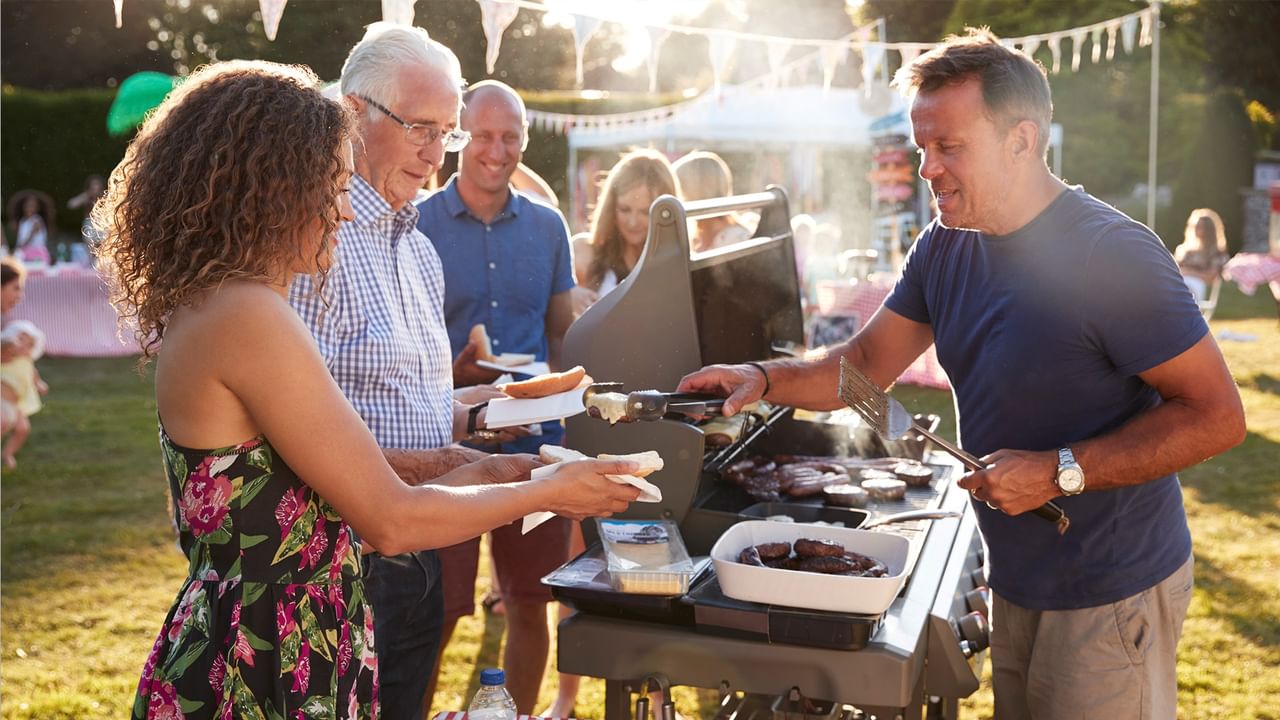 A group of people are gathered around a grill, with a man cooking and a woman handing him food.