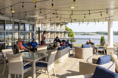 Guests relaxing on a patio deck under hanging string lights near a calm body of water at Rodd Brudenell River Resort