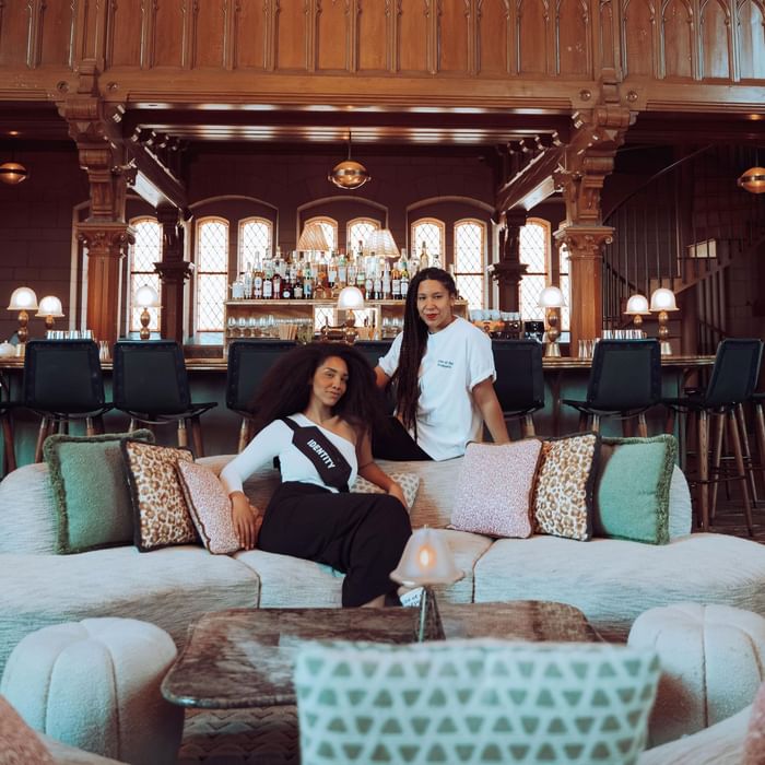 Leni Charles & Cherrie O posing on a plush sofa adorned with patterned pillows and shelves of drinks at Hotel Motto Vienna
