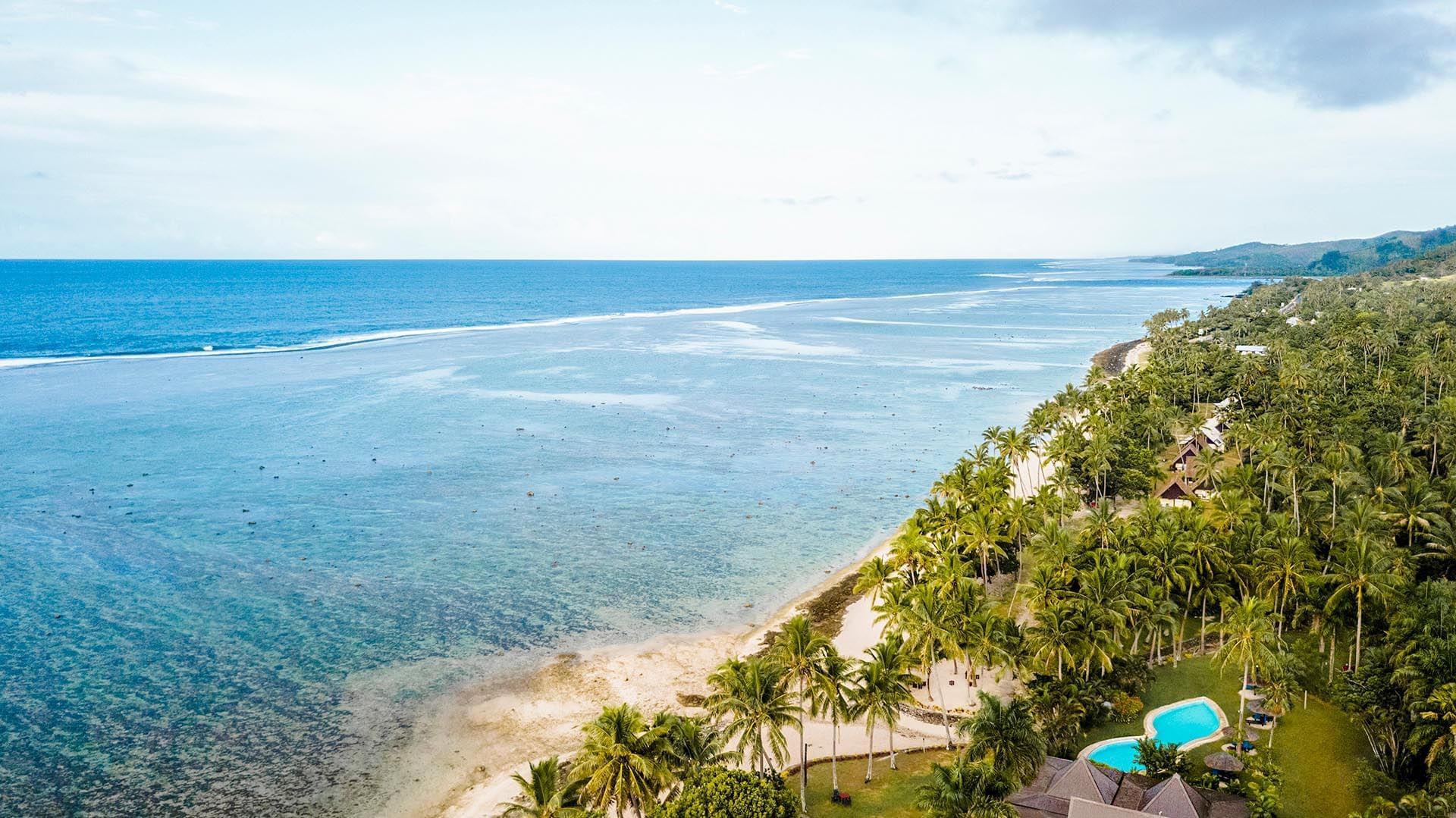 Wide aerial view of the coastline by a coral reef under a blue sky near the villas at Tambua Sands Beach Resort