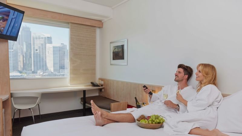 Couple relaxing in bathrobes on a bed at ibis Sydney Darling Harbour, with Sydney CBD skyline views