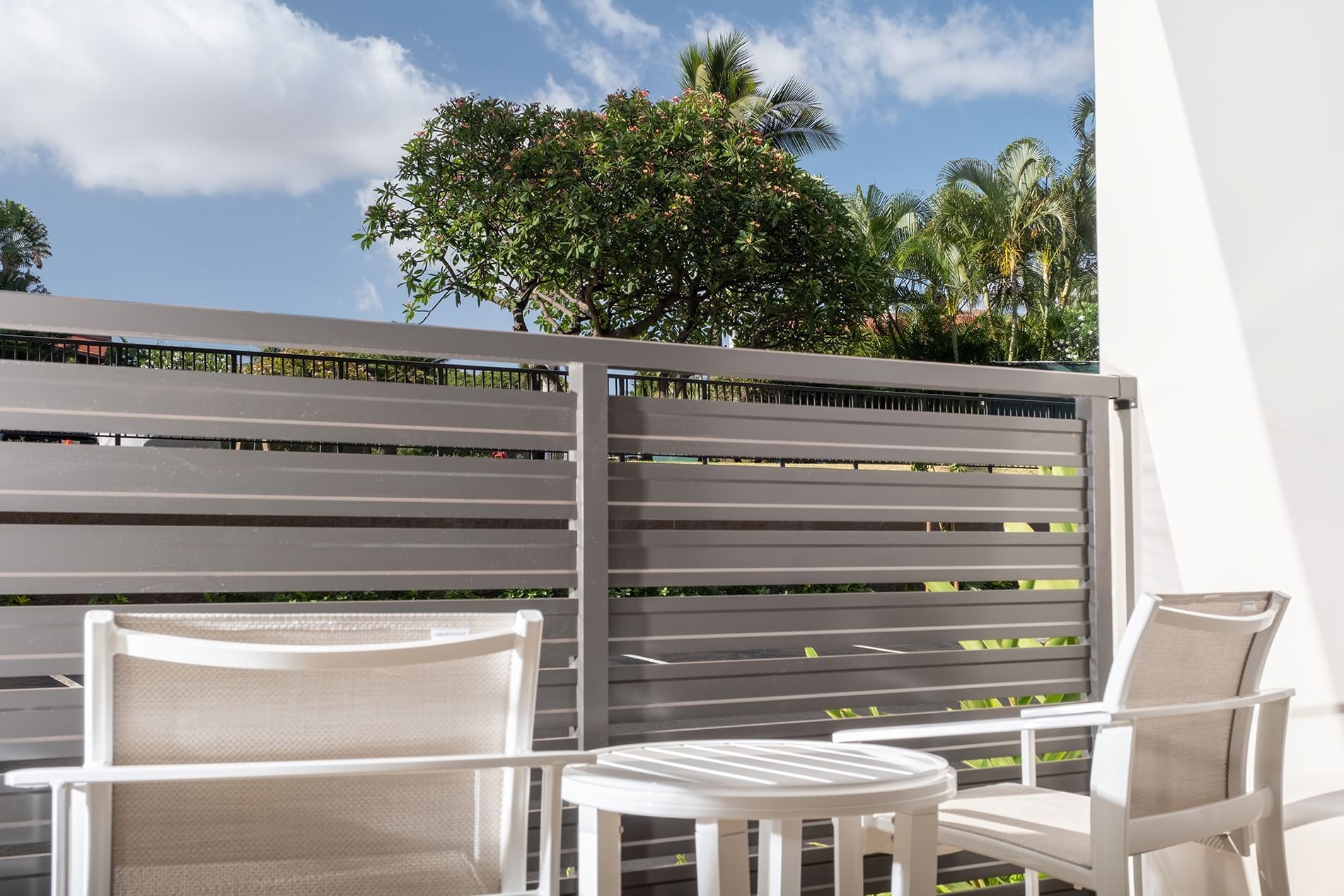A balcony with white furniture and a gray fence overlooking trees and blue sky.