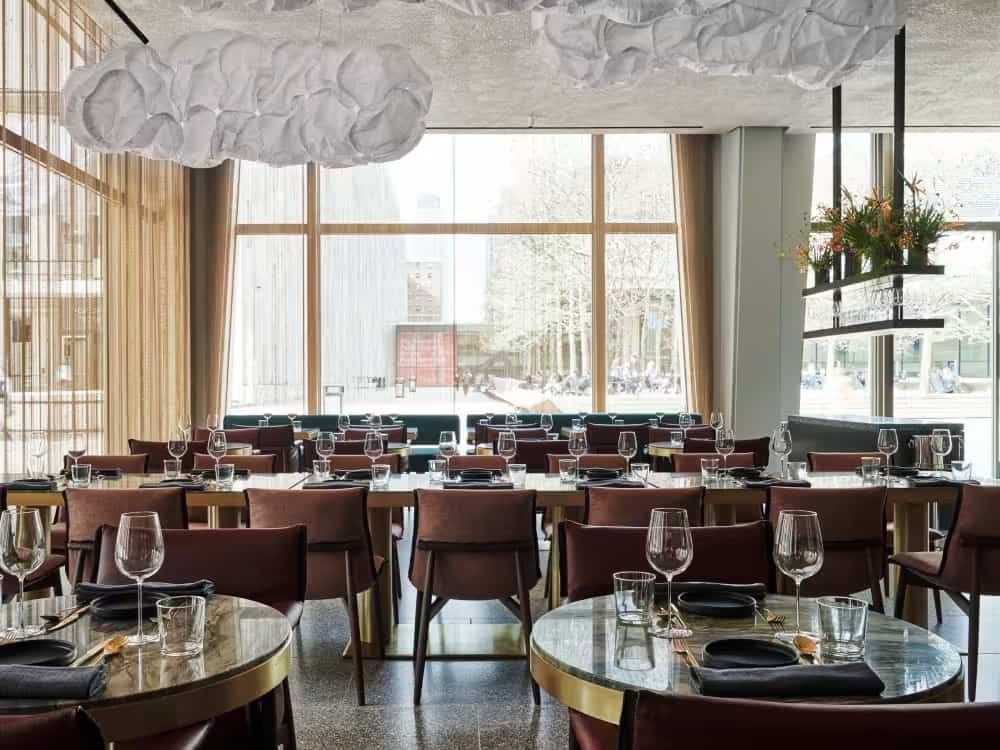 A dining room with round tables set for meals, large windows, and a unique cloud-shaped light fixture.