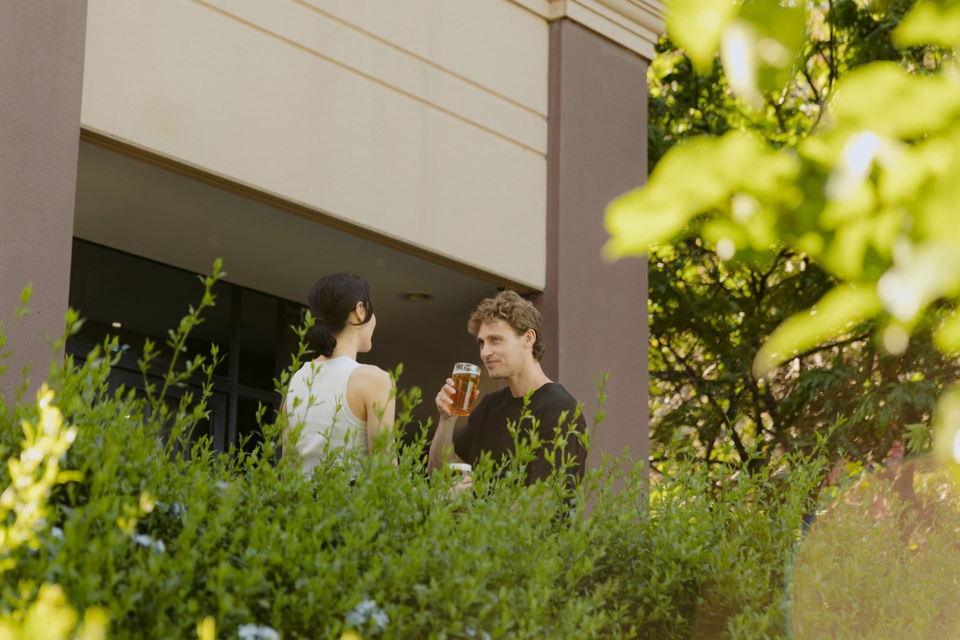Couple conversing outdoors by a building, surrounded by lush greenery at Amora Herencia Riverwalk Melbourne