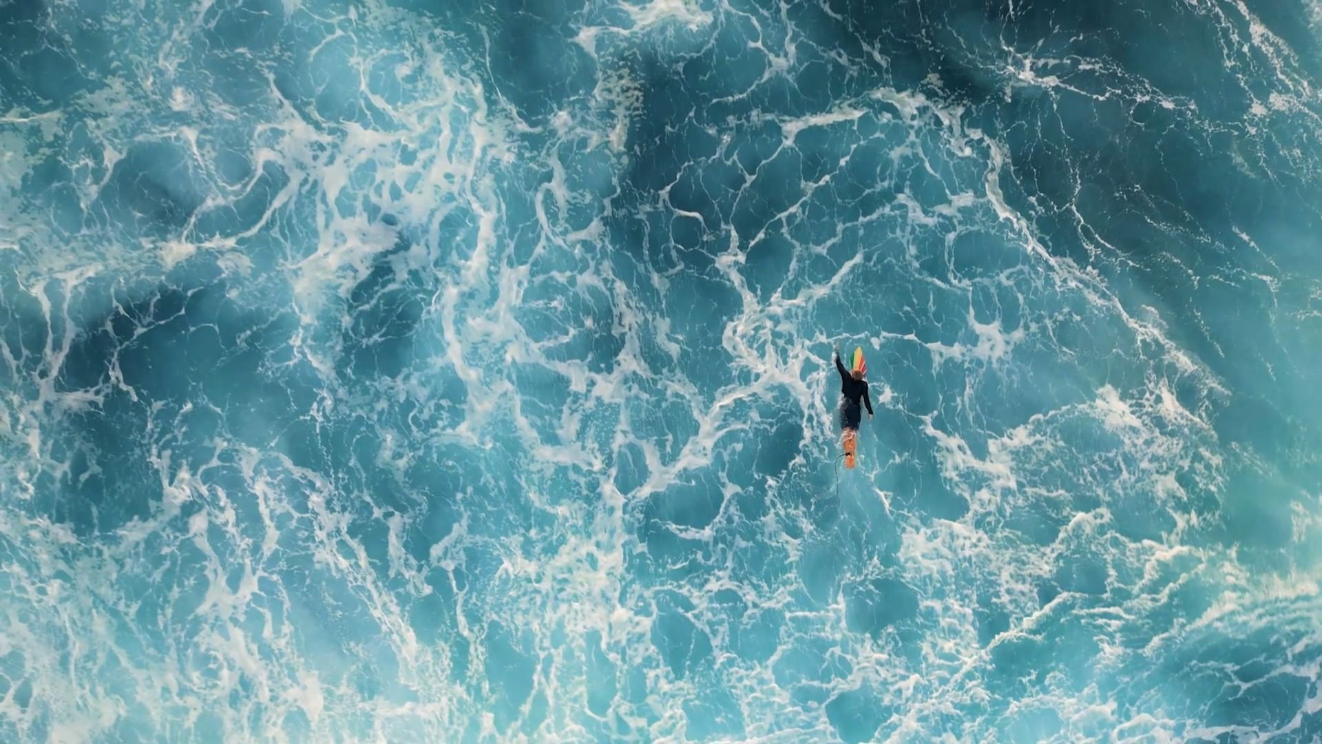 Aerial view of a surfer in black wetsuit holding a rainbow kite above turbulent blue ocean waves.